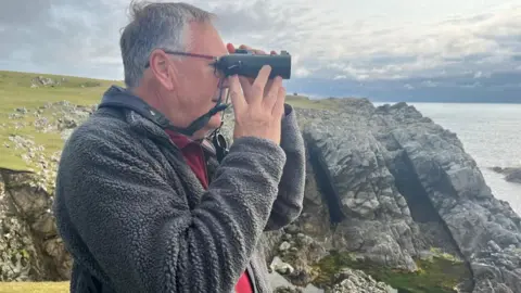 A man with greying hair and wearing a grey fleece jacket and red jumper. He is wearing glasses and holding binoculars to his eyes. He is standing on cliffs. More cliffs and the sea can be seen in the background.