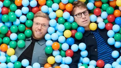 BBC Rob and Josh lie in a colourful ball pit, with their bodies mostly submerged. Their faces peer out and they are looking directly at the camera.