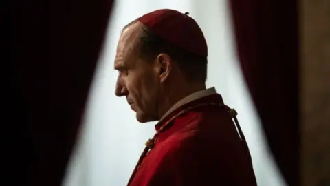 Ralph Fiennes standing side on in his role as a cardinal in the film Conclave. He is wearing red robes and a skull cap. Behind him is a window with parted curtains. He is closing his eyes in prayer.