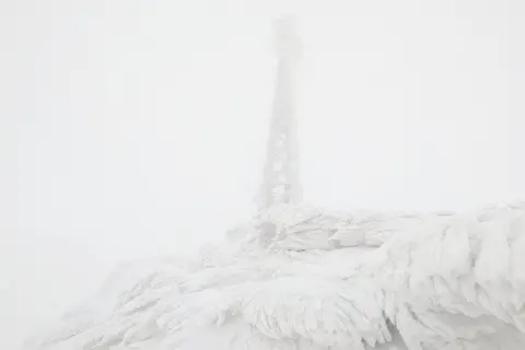 SAIS Glencoe The objects in the image are almost obscured by low cloud. The mast is caked in ice and rises above an area of ground covered in a meringue-like covering of ice.