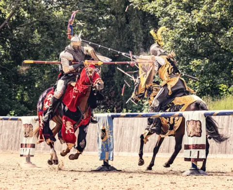 LAS Two men kitted in medieval metal armoury riding horses and charging at each other while holding long decorative poles. The horses are covered in elaborate cloths.