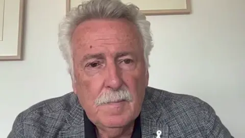 Prof Tim Chapman has grey hair and a grey moustache and is sitting in front of a white wall. He is wearing a grey checked suit jacket and black shirt.