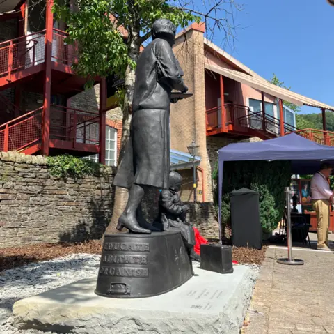 The back of a grey statue of a woman, who is wearing a skirt and heeled shoes. There is weiting on the plynth she is stood on which reads "educate, agitate, organise". behind the statue is a blue marquee with people stood under it.