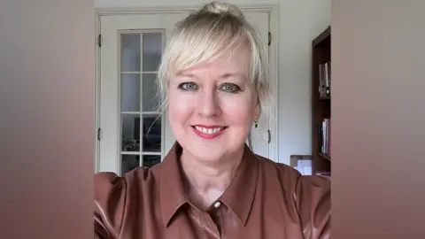 Dr Tanya Horek A woman takes a selfie and smiles. She has light blonde hair which has been tied back into a bun and she has a fringe that rests over her forehead. She is wearing a brown leather top. 