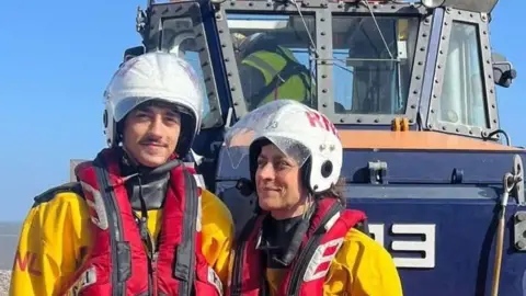 RNLI Minehead Both wearing yellow waterproof coats and red life-jackets and white helmets, Harrison stands on the left while his mum stands next to him, looking at him. Behind them is an RNLI craft.