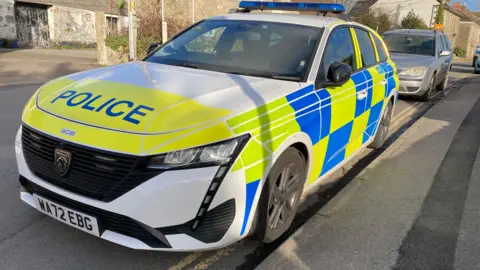 A white, blue and yellow Peugeot police car from Devon and Cornwall Police parked on a residential street in Cornwall. It's licence plate is WA72 EBG. No-one appears to be sat inside. A silver Ford saloon car is parked behind the police car.