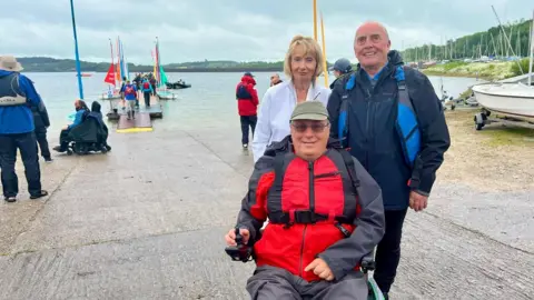 Two men and a woman are near a jetty filled with sailing boats on a lake. The man on the right has a blue life jacket, dark clothing and no hair. The man in the centre has a red life jacket, a dark coat, green cap, sunglasses and is sat in an electric wheelchair. The woman has short blonde hair and is dressed in a white jacket. There are people walking in the distance and one man in an electric wheelchair.