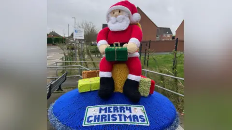 Syston Knitting Banxy A knitted postbox topper depicting Santa Clause sitting on a chair surrounded my gifts with a blue base that says 'Merry Christmas' 