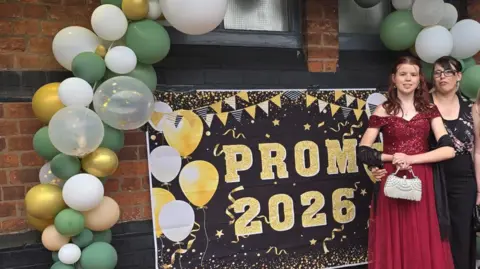 Lillie-Mae is standing to the right of the picture in front of a black sign with the words 'Prom 2026' in gold lettering. The sign also has white and gold balloons and bunting on it. Lillie-Mae is wearing a red off the shoulder dress with a black shawl draped over both arms. She is carrying a white handbag. To her left is her mum Katie, who is wearing a black top with a multi-coloured pattern.