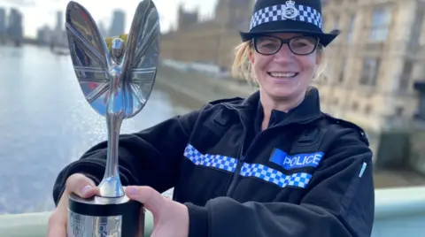 Martis Media A police officer in her uniform, holding a silver trophy with a silver winged figure on top