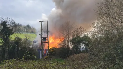 A fire can be seen in the distance across a steel metal fence. There is a large smoke cloud.