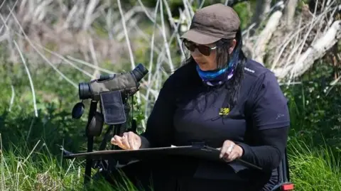 Melanie Mascarenhas is wearing a brown hair, dark sunglasses and black top.
In her lap is a large painting of the green field that she is sitting in. 
