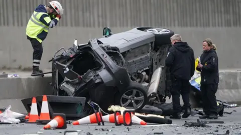 PA Media Three crash investigators examine an overturned car. It is mangled. Several red-and-white traffic cones are scattered nearby.