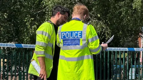 Two police officers. wearing high-vis police jackets, have their backs to us as they talk while reading a document. They are standing in front of blue and white police tape stretched across a road.