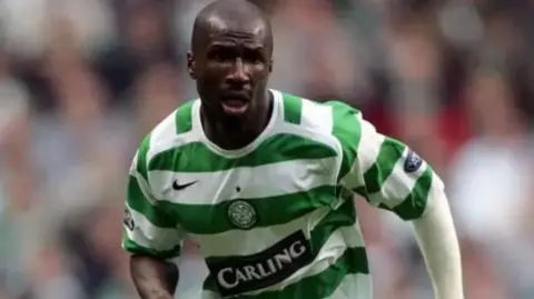 Camara is wearing a Celtic kit and has an arm bandage on and is captured playing during a match