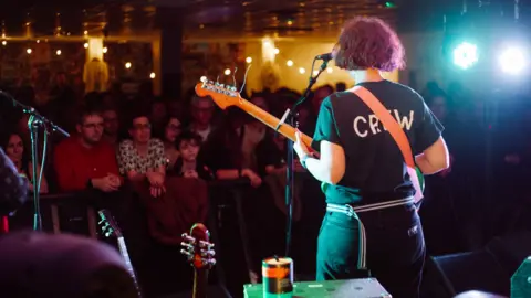 A person on stage, pictured from behind. They are wearing black and are holding a guitar, with a microphone positioned in front of their face. In the background is a standing audience watching on. It is dimly lit, with stage lighting.
