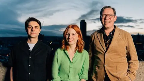Three people stand on a balcony with a cityscape at dusk behind them. Dino has straight black hair and wears a black buttoned up shirt. Alice has ginger hair and a bright green buttoned-up shirt. O'Hara is significantly taller than the other founders and weas 