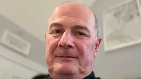 A bald man looks into the camera. Behind him are paintings hanging on his wall 