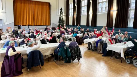 Rows of tables with people sat around them in a hall. Some people have their hands up in the air waving at the camera, some are just smiling. There are around eight tables with white table clothes on them. Some people are wearing paper hats from crackers. 