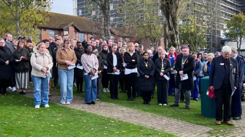 A crowd of people standing with heads bowed in front of the large hospital tower block