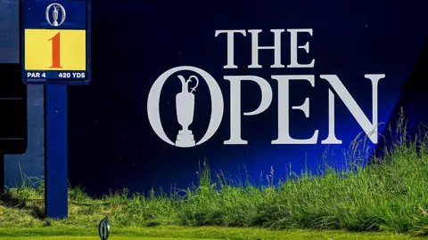 PA Media A large blue sign with the Open branding. The words are in white font and the claret jug is in the centre of the O. A signpost for the first tee is to the left of the picture. It has a large red digit one on a yellow background.