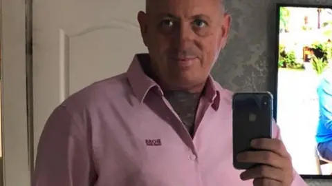 Facebook A middle aged man takes a selfie with his mobile phone in a bedroom mirror. He is wearing a pink Hugo Boss branded buttoned shirt with an open collar, has short grey hair and the front of his chest up to his neck is covered in tattoos. 