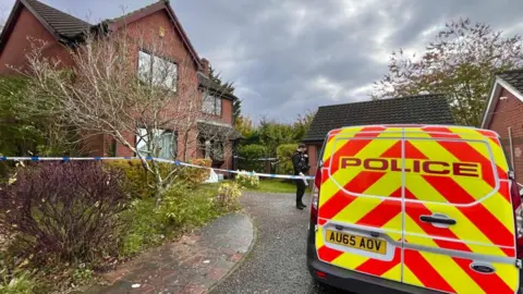 A police forensic van is outside a house in Norwich. There is a cordon outside the modern-looking which has two garages. The property is well-kept with lawns and hedging. A police officer is standing outside the house, on the road.   