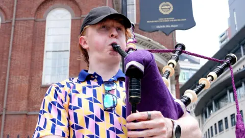 Piper playing bagpipes in front of a historic red-brick building near a sign reading 'Boston Common Founded 1634', with a clock tower and city street scene in the background