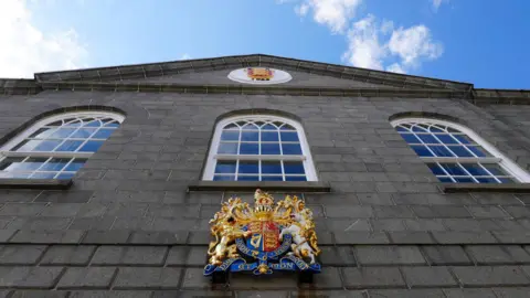 BBC A view of the outside of the Guernsey States chamber showing a large gold coloured coat of arms and windows. 