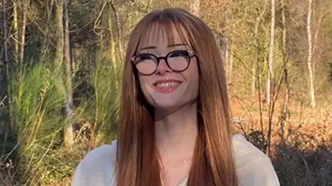 Family photo Brianna Ghey, with long red hair and glasses, standing with forestry behind her 