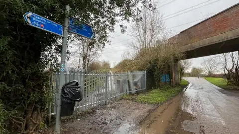 A view of an entrance to the greenway on Holmes Lane, showing metal fencing blocking the path.