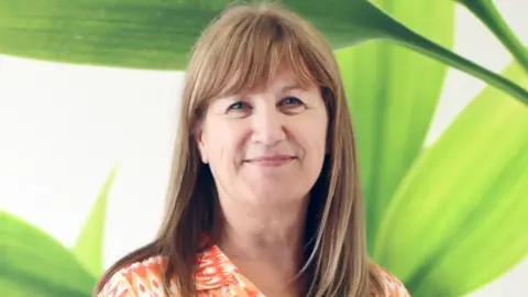 Farleigh Hospice A woman with brown shoulder-length smiling at the camera with a leafy green wallpaper in the background.