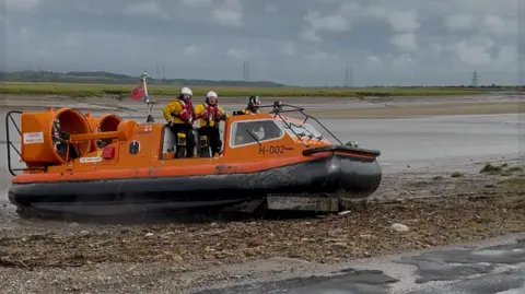 Two lifeboat crew on the deck of their hovercraft by the river