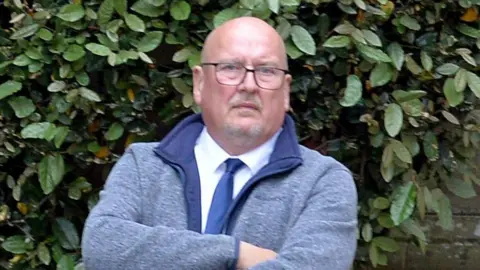 A bald-headed man with glasses and a goatee beard in a blue fleece top with white shirt and blue tie underneath. There is a hedge behind him.