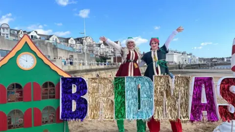 Bridmas day celebration with two stilt walkers dressed as elves on Bridlington beach standing next to a green, wooden house in front of a tinsel Bridmas sign 