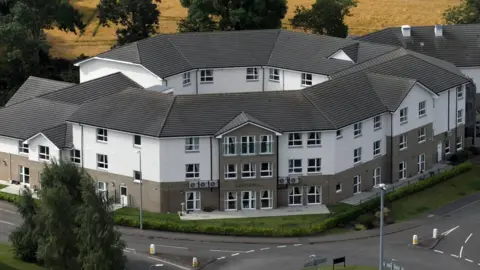 An exterior shot of a large modern care home. It is a grey and white building with three floors and rows of windows in its walls. There is a road with a roundabout near the main entrance.