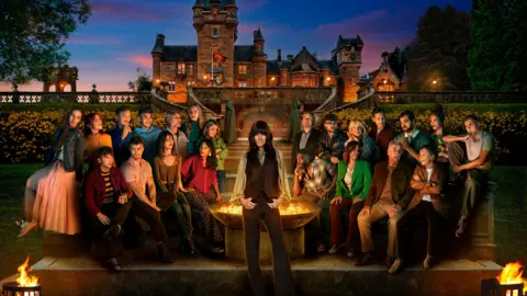 Cody Burridge/BBC A press photo of the contestants of series four of The Traitors, sitting around a fire outside a castle. Host Claudia Winkleman stands in the centre, smiling at the camera.