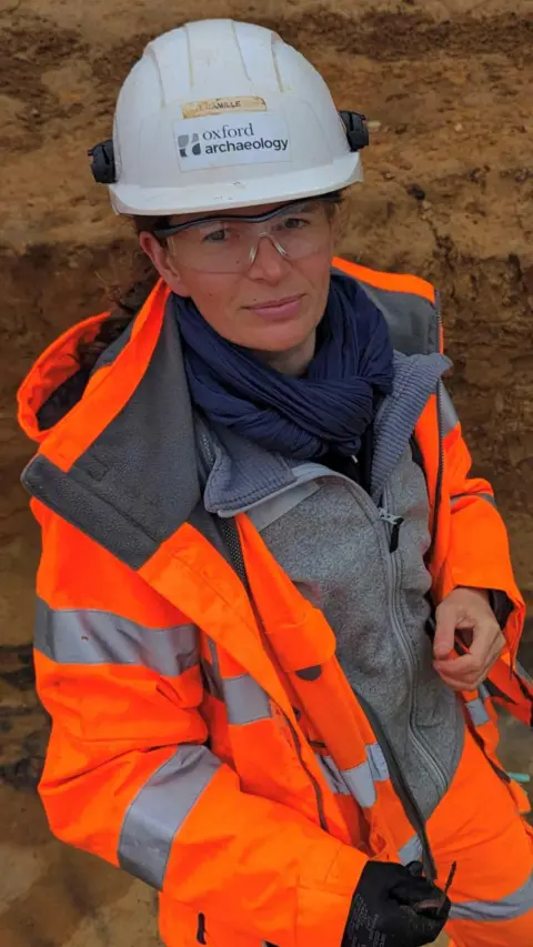 Oxford Archaeology Camille Guezennec with white hard hat, safety glasses, wearing an orange hi-vis jacket, grey fleece, blue scarf and black gloves. She is on a burial site.