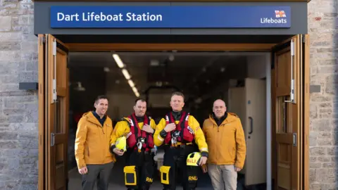 A picture of four men stood out the front of the lifeboat station. There is a sign above the door that reads "Dart Lifeboat Station". There is a person each side wearing a yellow coat. There is two people in the middle wearing a yellow jacket and a lifejacket.