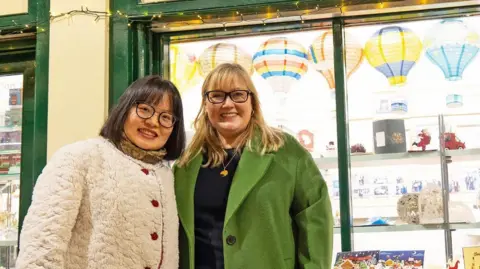 Karen Kilgour with the owner of Tian's Gifts in Grainger Market. Kilgour has shoulder-length blonde hair and is wearing a green coat. The stall owner is a woman with a black bob cut and glasses. She is wearing a thick cream woollen coat with red buttons. They stand in front of a gift shop with decorative miniature hot air balloons in the window.