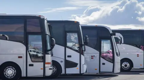 Four white coaches are parked alongside one another in a car park.
