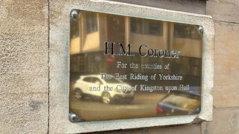 A brass plaque on the wall outside Hull Coroner's Court, with cars reflected in it. It reads "HM Coroner for the counties of the East Riding of Yorkshire and the City of Kingston upon Hull"