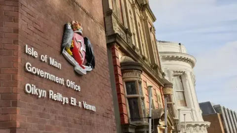 BBC Isle of Man government offices, which has lettering on a red brick wall. The wedding cake Tynwald Building can be seen along side it.