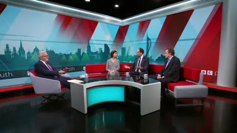 A wider view of the studio, revealing presenter Peter Henley who is interviewing the three guests.