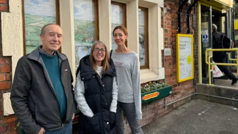 Tom Gillespie, met een grijze fleece en turquoise trui, Marie Gillespie met een donkerblauw gilet en lichtgrijze trui en Kerry Baynes, met een lichtgrijs sweatshirt en een spijkerbroek. 