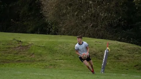 Spencer Wadsworth Spencer is on a green rugby pitch next to a corner post. He has the rugby ball in his hands and is running  forwards to take a kick. He is wearing a white top and black shorts.