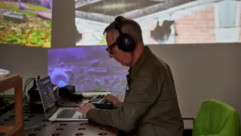 Judy Hume Steve Jones is working on a laptop at a desk with black headphones on and a green coat over his chair. There are four projector images on the wall behind him.