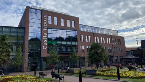 A modern-looking office building, with formal gardens in front of it. There are paved walkways and a bandstand, with people sitting on benches dotted around the space in front of the building. There is lettering on the side of the building which says Castle House.
