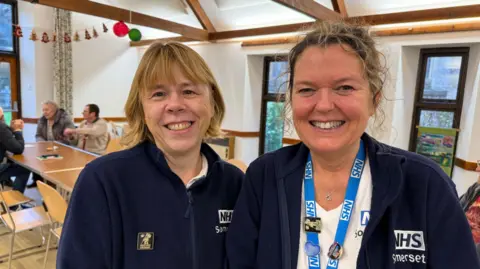 Lady with light, shoulder-length hair smiles at the camera to the left of her NHS colleague with curly hair wearing a blue lanyard who also smiles at camera.