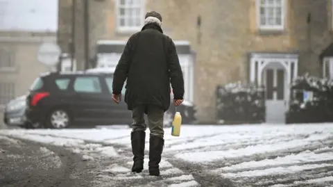 A man in boots and a jacket is seen with his back to camera, holding a carton of milk. He is walking across snow. 